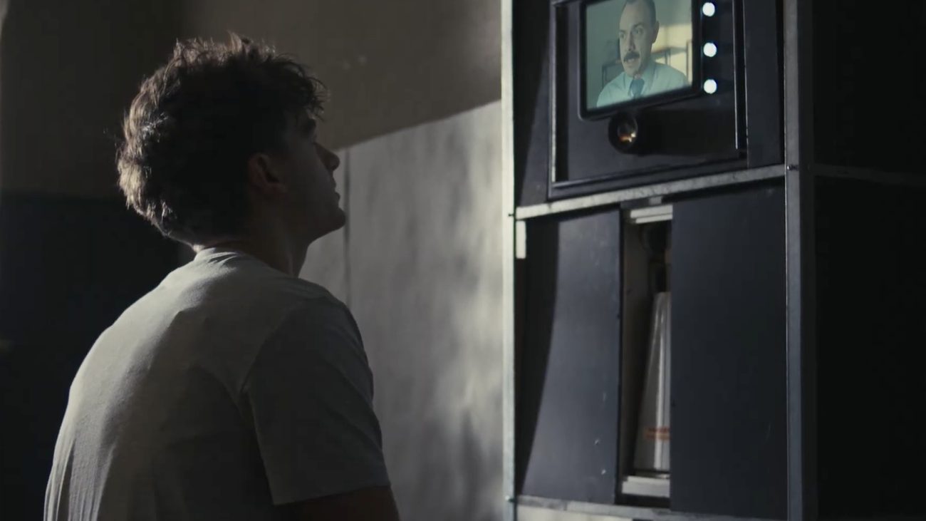 Film still: a man seen from behind looking at a small screen